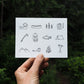 a greeting card featuring small line illustrations of camp and adventure things; tents, a backpack, a fish, a fishing rod, mountains, a mug and kettle, a canoe, a campfire, marshmallows and hotdogs on sticks, an axe and a field book. The designs are on a nice textured white paper resembling watercolour paper.